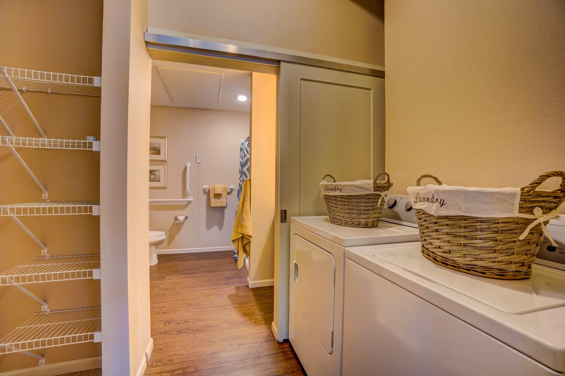 Laundry area with a washing machine and dryer, each topped with a wicker laundry basket labeled 'Laundry'. To the left, an open doorway leads to a bathroom with a toilet, grab bars, and a towel hanging on the wall. The floor is wood, and there are wire shelves on the left side of the laundry area.
