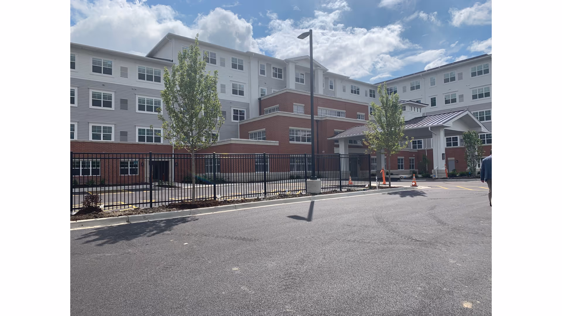 Exterior view of a multi-story senior living facility building with a combination of brick and light-colored siding. The building has many windows and a covered entrance. There are young trees planted along the sidewalk and a black metal fence in front. The sky is partly cloudy.