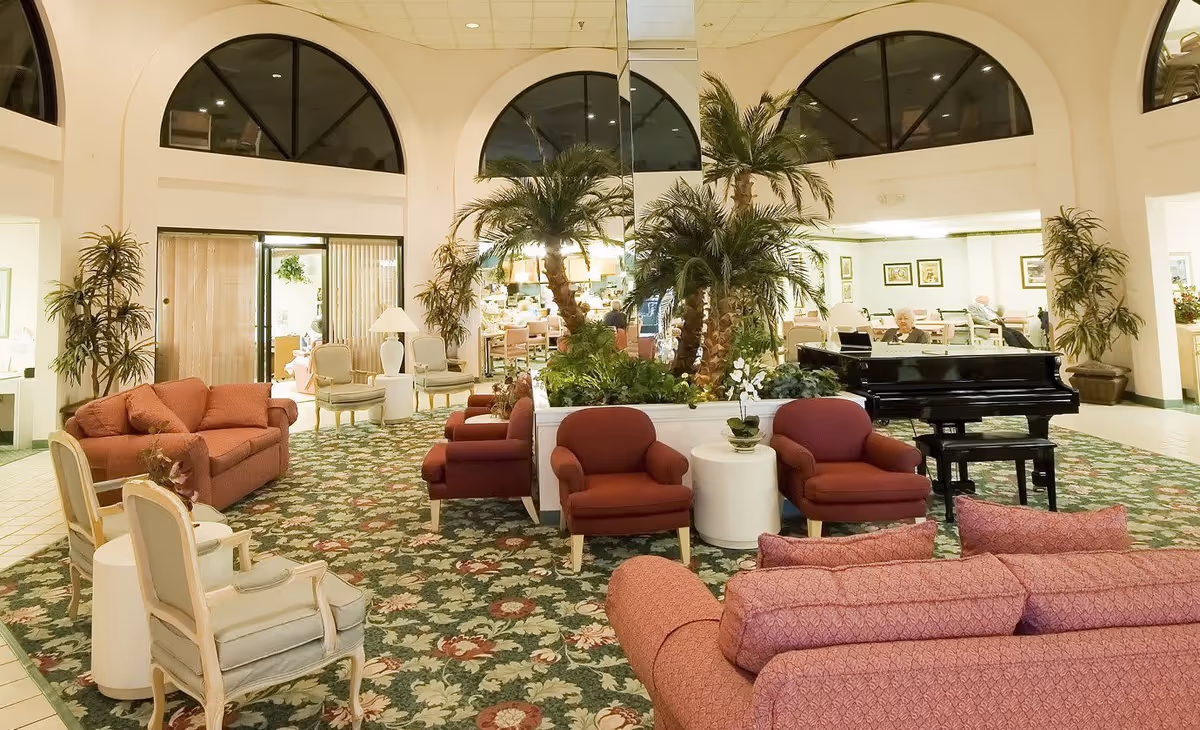 A spacious and well-lit senior living facility common area with floral carpet, multiple seating arrangements including red and beige armchairs and sofas, a black grand piano, and large potted plants. The room has high ceilings with arched windows and a mirrored wall behind the plants. In the background, elderly residents are seated at tables.