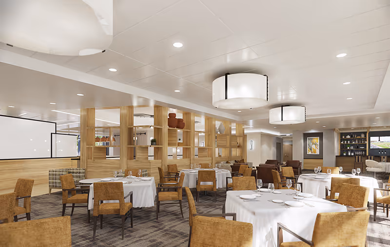 A spacious dining room with round tables covered in white tablecloths, set with plates, glasses, and silverware. The room features brown upholstered chairs, modern ceiling lights, wooden partition shelves with decorative items, and a bar area in the background.