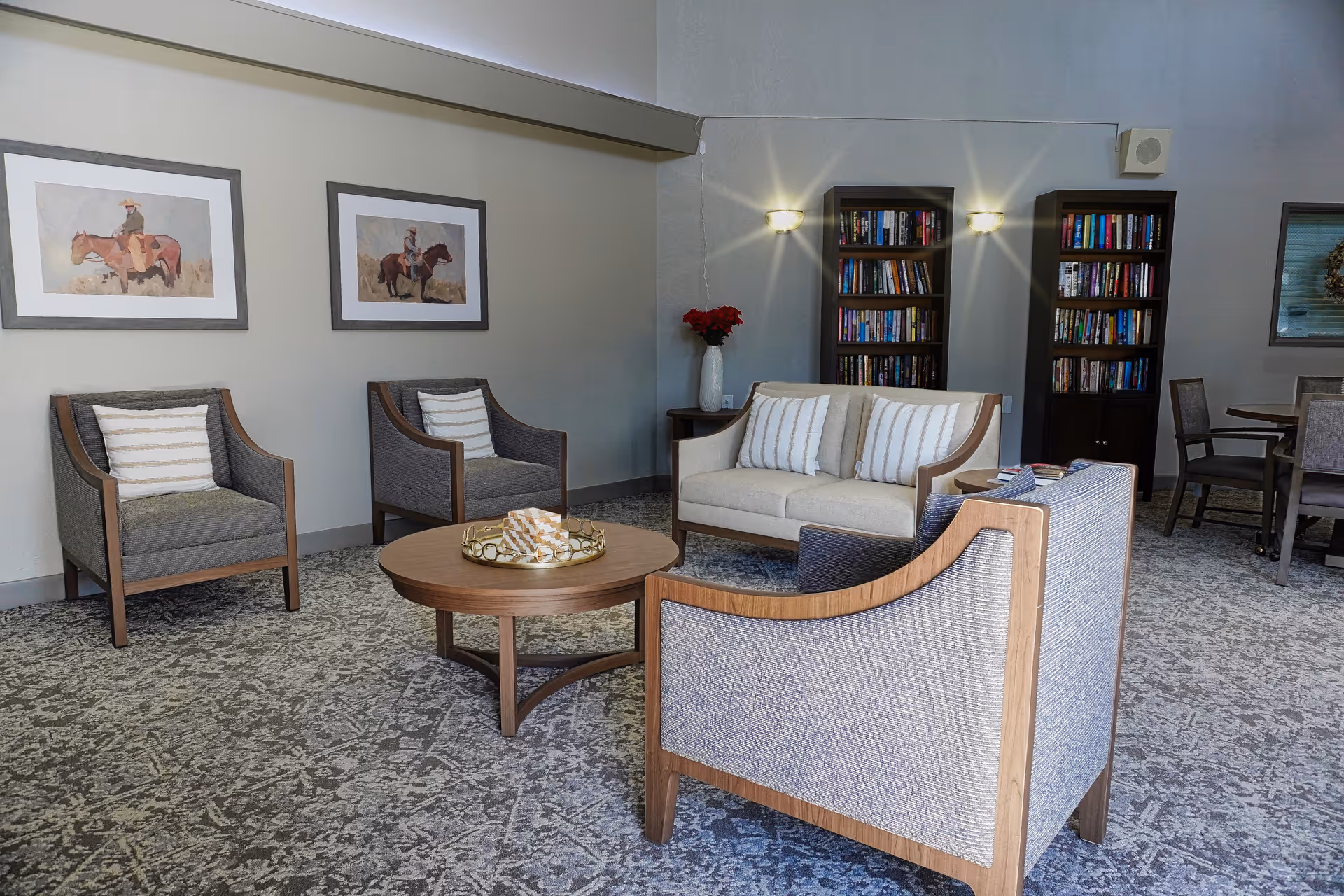 A cozy living room area with a beige loveseat and three gray armchairs arranged around a round wooden coffee table. The coffee table has a decorative tray with a folded cloth. Behind the loveseat are two tall bookshelves filled with books and two wall-mounted lights above them. On the left wall, there are two framed pictures of people riding horses. To the right, there is a small dining area with a round table and chairs. The room has a patterned carpet and neutral-colored walls.