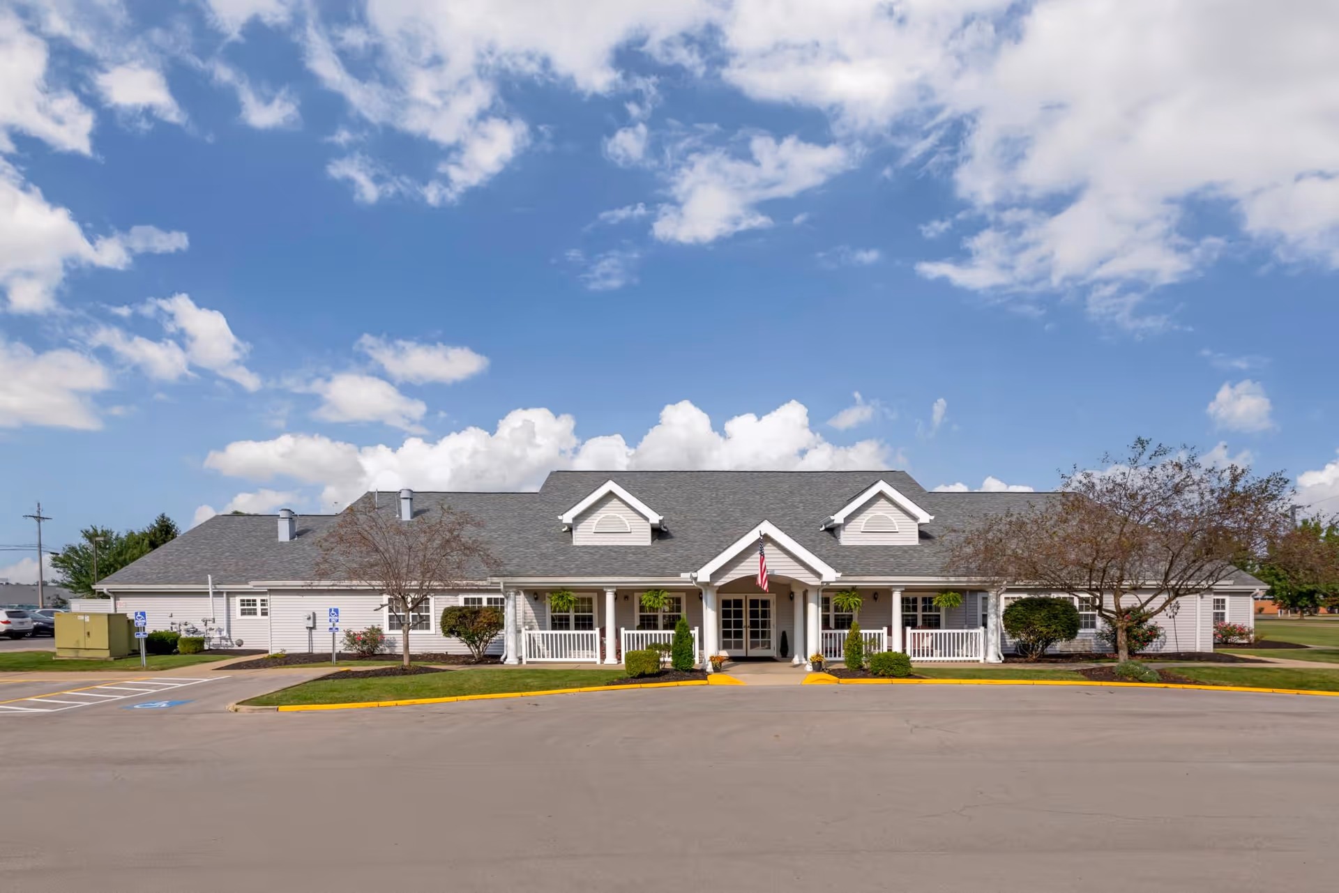 Front exterior view of The Addison of Garden Way Place, a single-story building with a gray roof, white siding, and a covered entrance with an American flag. The building is surrounded by a paved driveway, landscaped bushes, and trees under a partly cloudy blue sky.