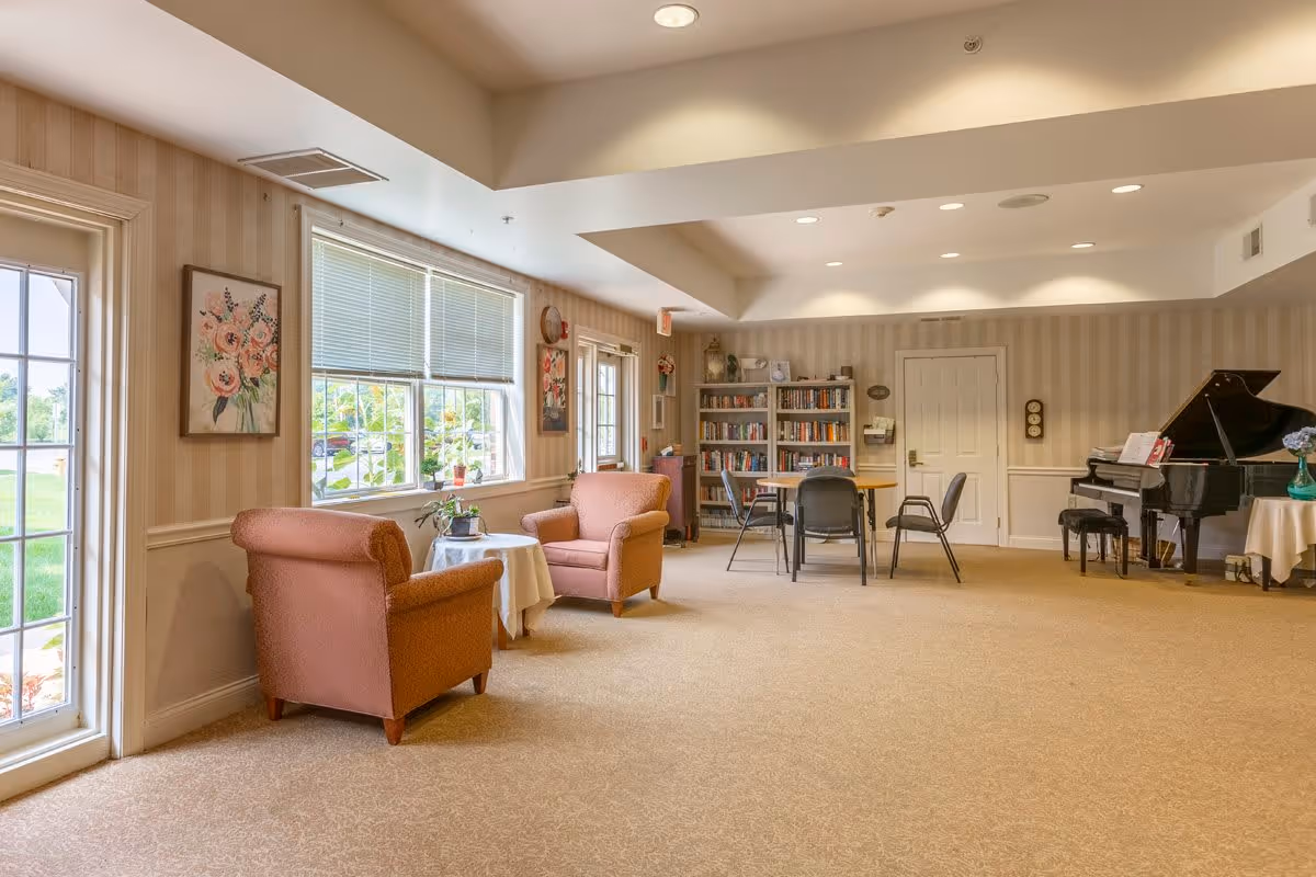 A cozy common area with two pink armchairs around a small round table near a window, a bookshelf filled with books, a table with chairs, and a black grand piano in the corner. The room has beige carpet, striped wallpaper, and soft lighting from recessed ceiling lights.