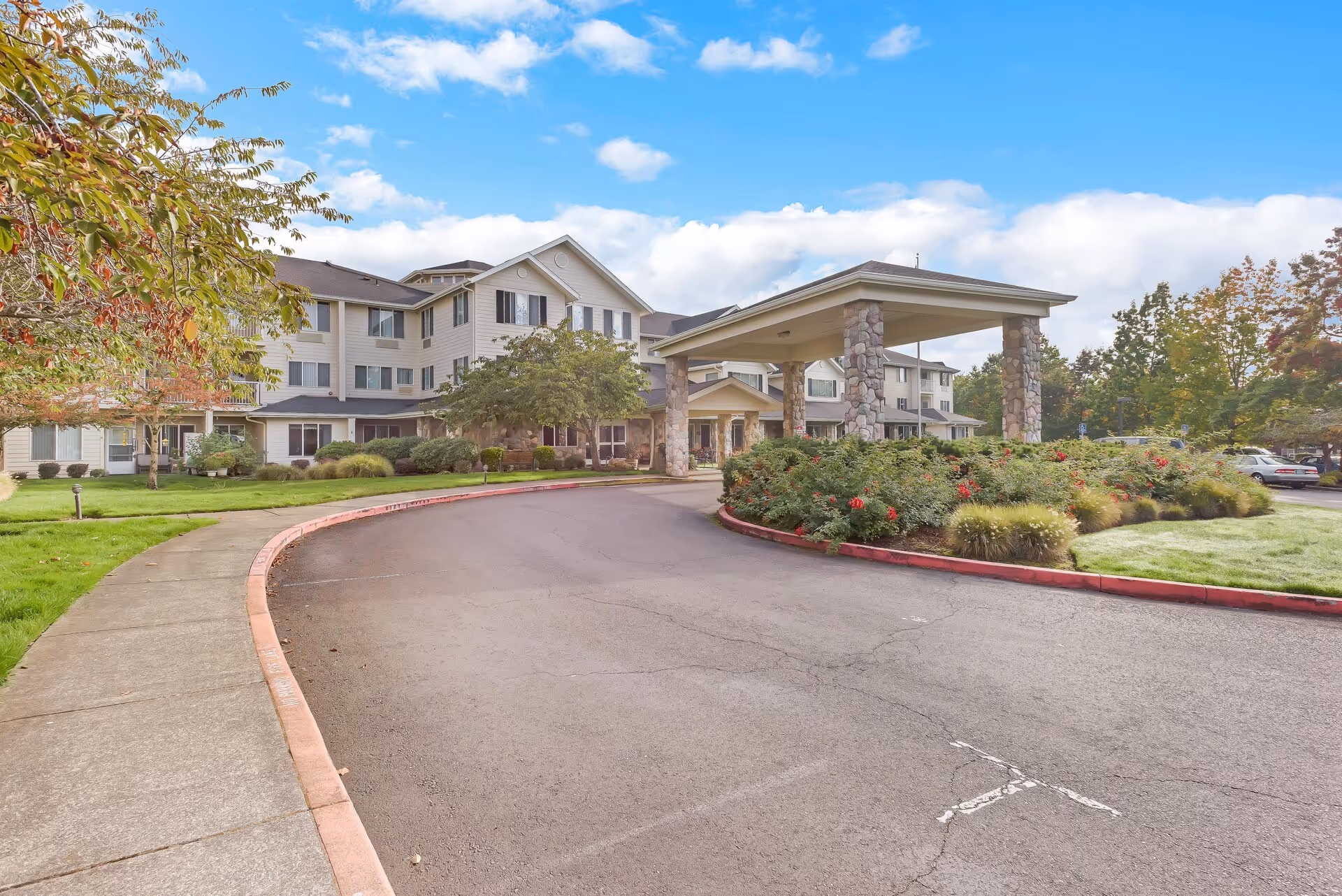 Exterior view of Solista Newberg senior living facility showing a three-story building with beige siding and dark shutters. There is a covered entrance supported by stone pillars, a curved driveway, landscaped bushes and trees, and a partly cloudy sky.