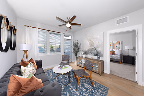 A bright and modern living room with a gray sofa adorned with brown and patterned pillows, two woven chairs around a round marble coffee table on a blue patterned rug. A wooden sideboard with decorative items and a large abstract painting is against the wall. A ceiling fan with lights is mounted on the white ceiling, and large windows with white curtains allow natural light to fill the room. An open doorway leads to a bedroom with a bed and nightstand visible.