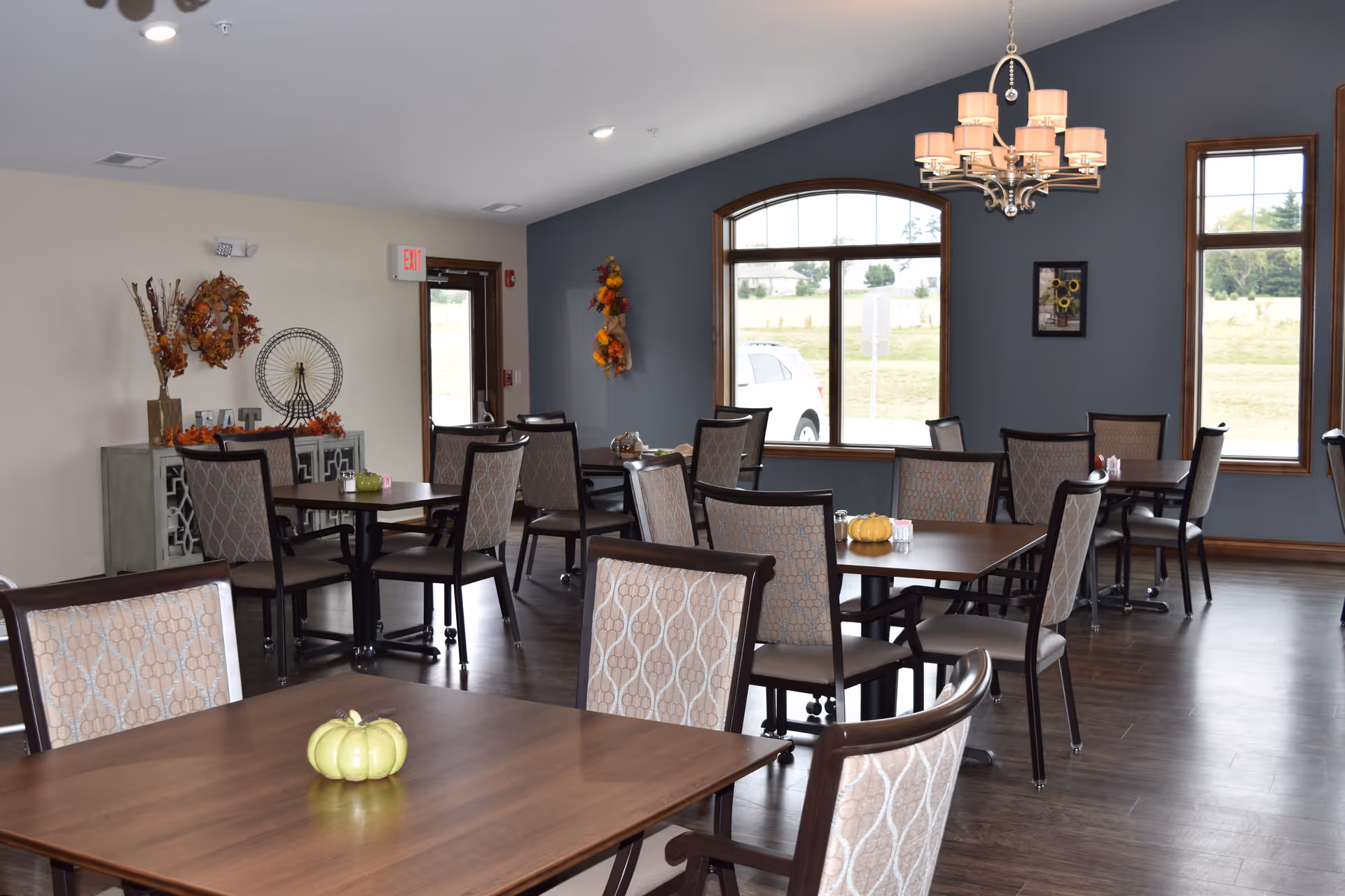 A senior living facility dining room with multiple wooden tables and cushioned chairs arranged neatly. The room has large windows letting in natural light, a chandelier hanging from the ceiling, and autumn-themed decorations including small pumpkins on the tables and wreaths on the walls.