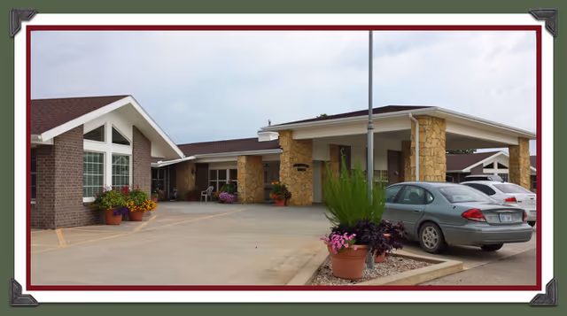 Exterior view of Moundridge Manor showing a single-story building with stone and brick facade, a covered entrance supported by stone pillars, several potted plants with flowers near the entrance, and a few parked cars in the driveway under a cloudy sky.