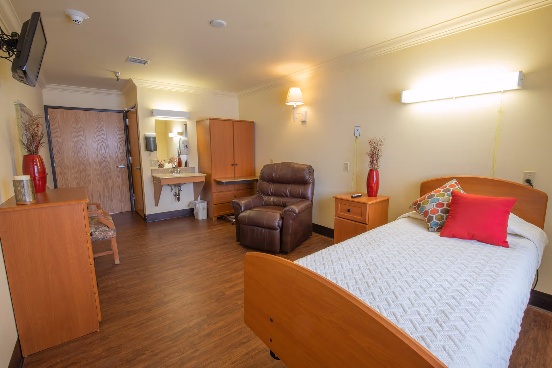 A bright assisted-living bedroom featuring a single bed with colorful pillows, a leather recliner, wooden furniture, and a small sink area.