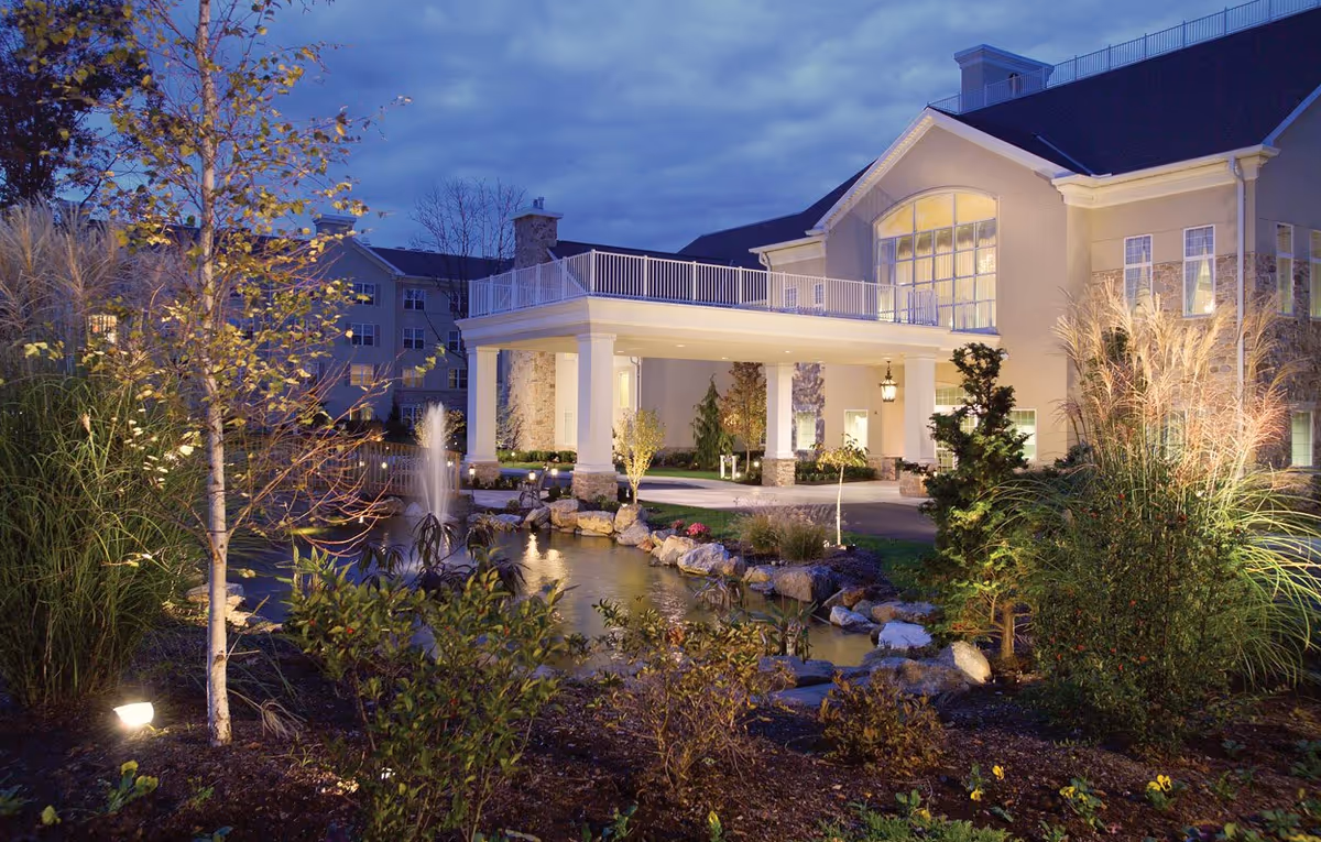 Evening view of the entrance to a senior living facility named Shannondell, featuring a covered driveway with white columns, a landscaped garden with various plants and bushes, a small pond with a fountain, and the building illuminated with warm lights under a cloudy sky.