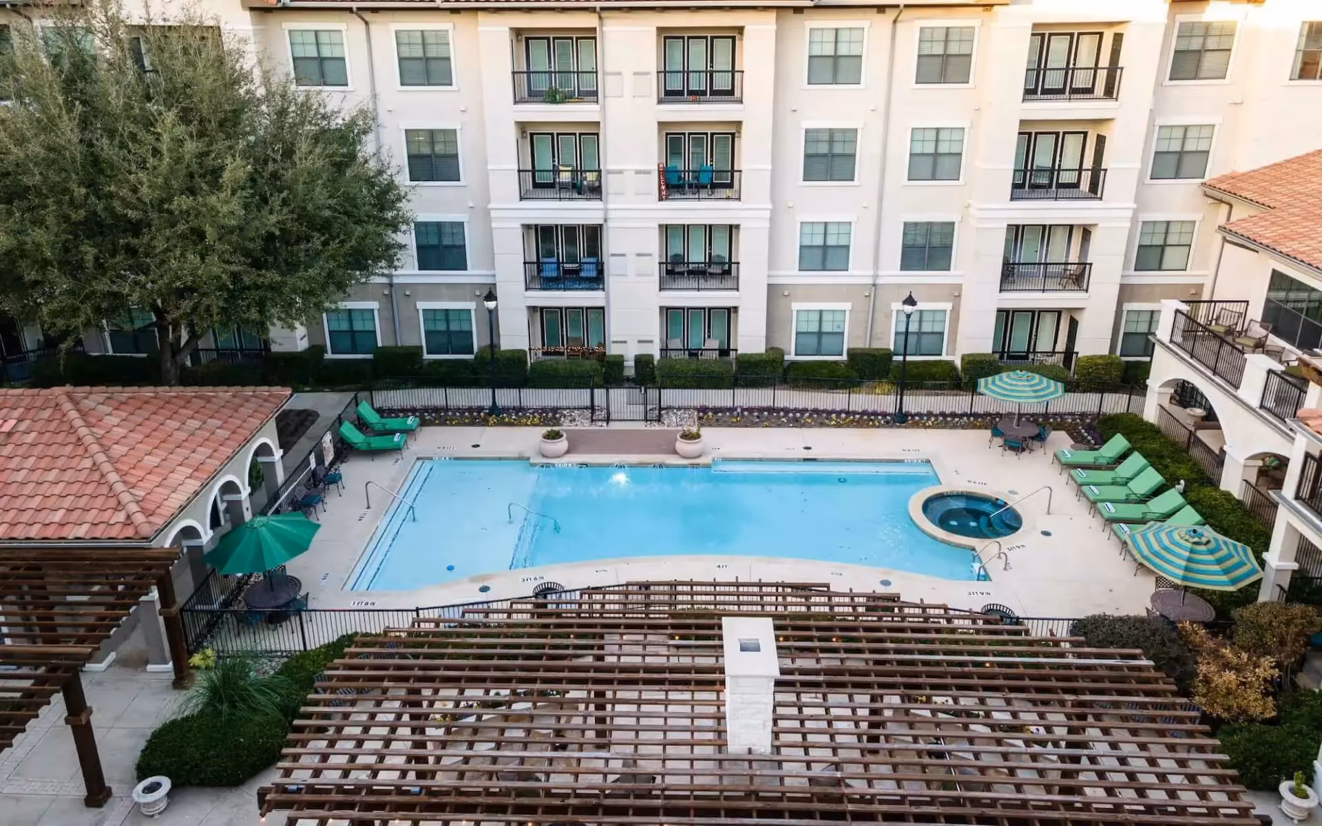 Aerial view of an outdoor swimming pool area at a senior living facility with lounge chairs, umbrellas, a hot tub, and a pergola. The pool is surrounded by a multi-story building with balconies and windows.