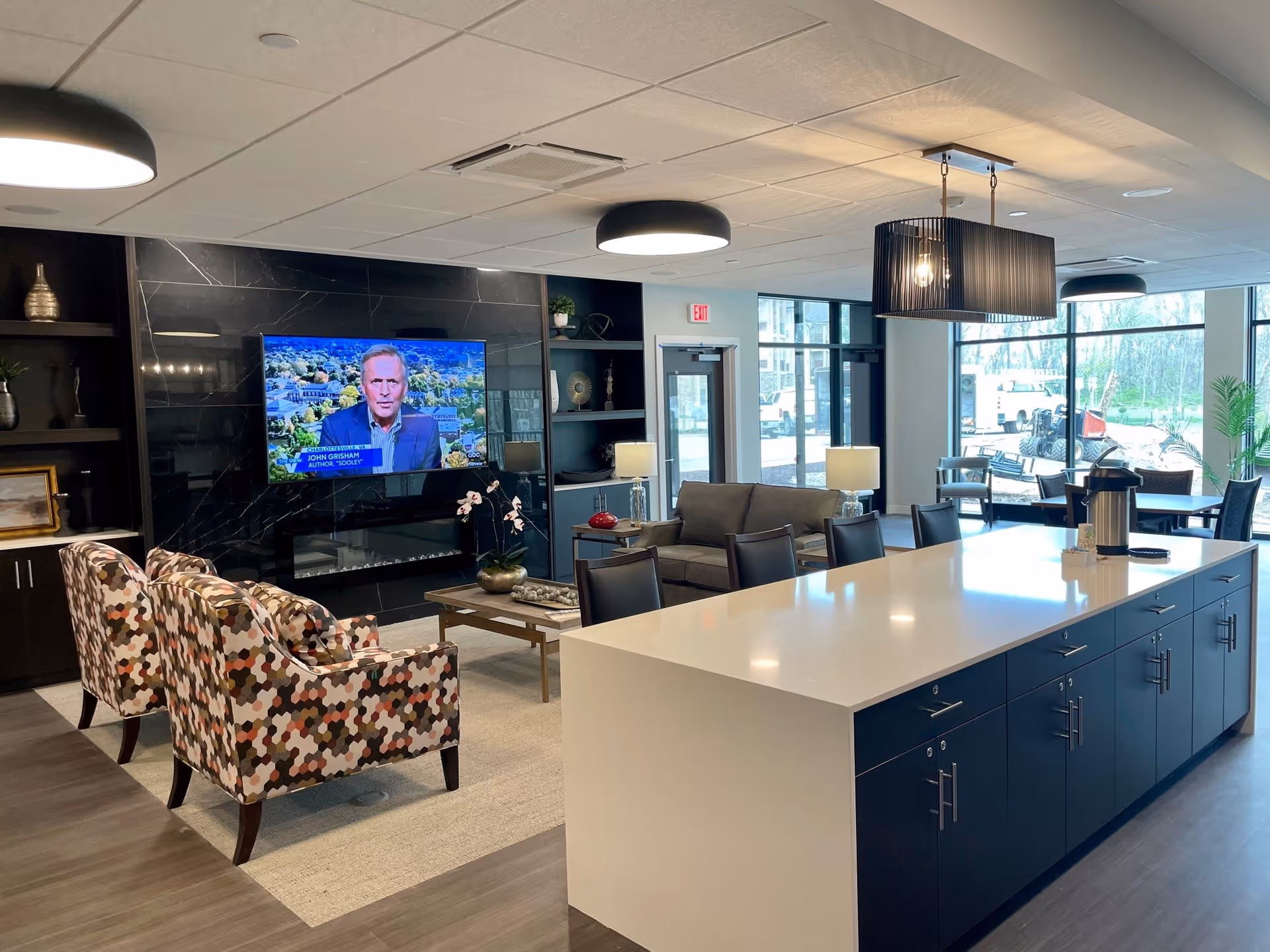 A modern common area with a large white island countertop with dark blue cabinets underneath, surrounded by chairs. In the background, there is a seating area with a gray sofa, two patterned armchairs, a coffee table, and a large flat-screen TV mounted on a black marble wall. The room has large windows letting in natural light and contemporary ceiling lights.