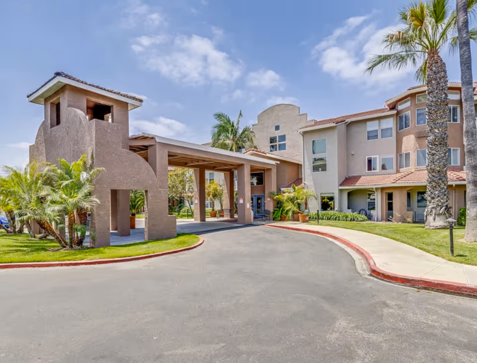 Exterior view of La Mirada Heights senior living facility showing the entrance with a covered driveway, palm trees, and a clear blue sky.
