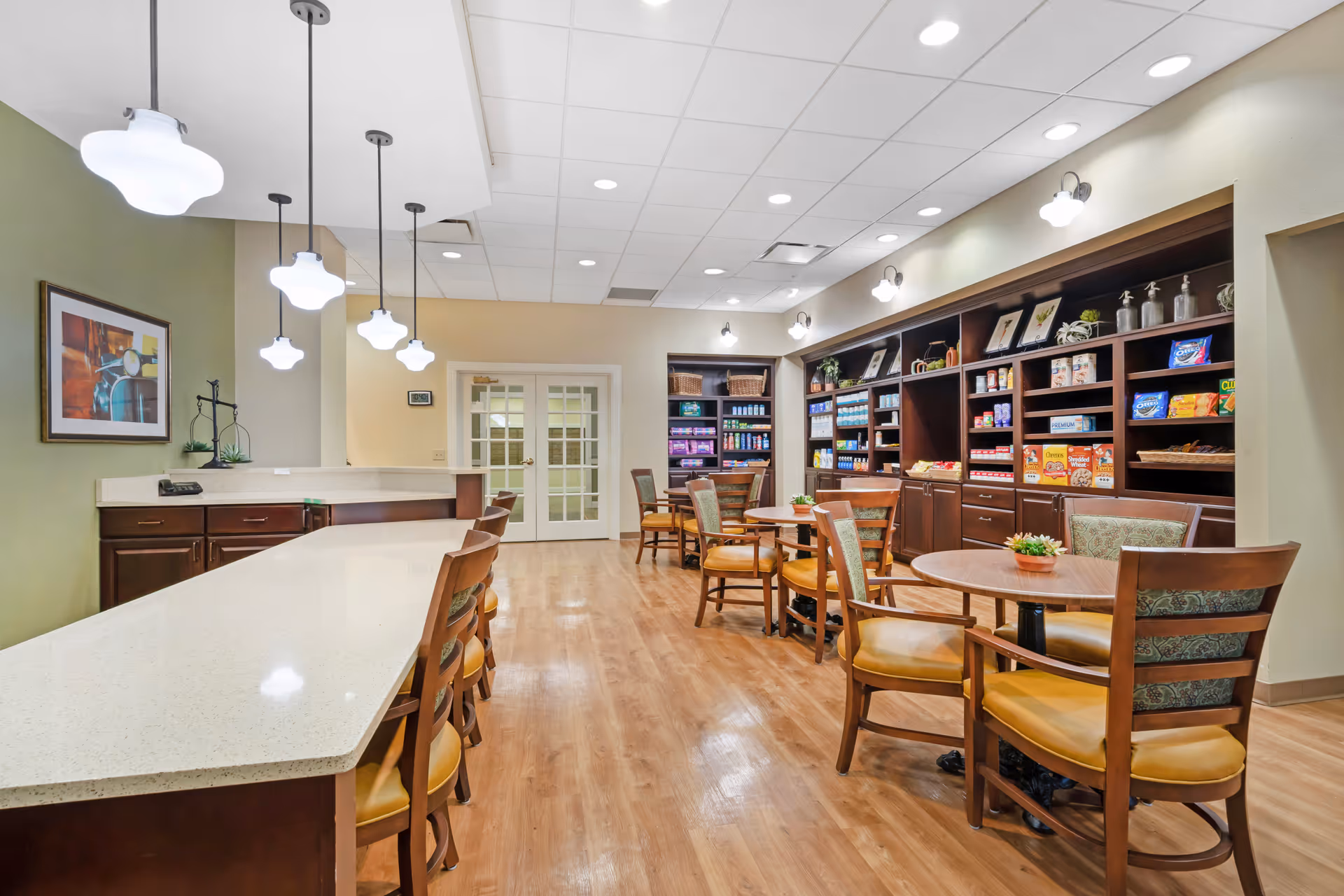 Interior of a senior living facility's common area with a long white countertop and several wooden chairs with yellow cushions. There are round wooden tables with chairs arranged around them, and shelves stocked with various snacks and items along the back wall. The room has wooden flooring, pendant lights hanging from the ceiling, and a framed picture on the wall.