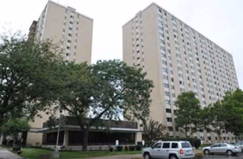 Two tall beige apartment towers with a front entrance, trees, and parked cars.