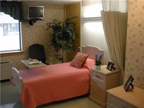 Private senior living bedroom with pink bedspread, nightstands, plant, window, and framed photos.