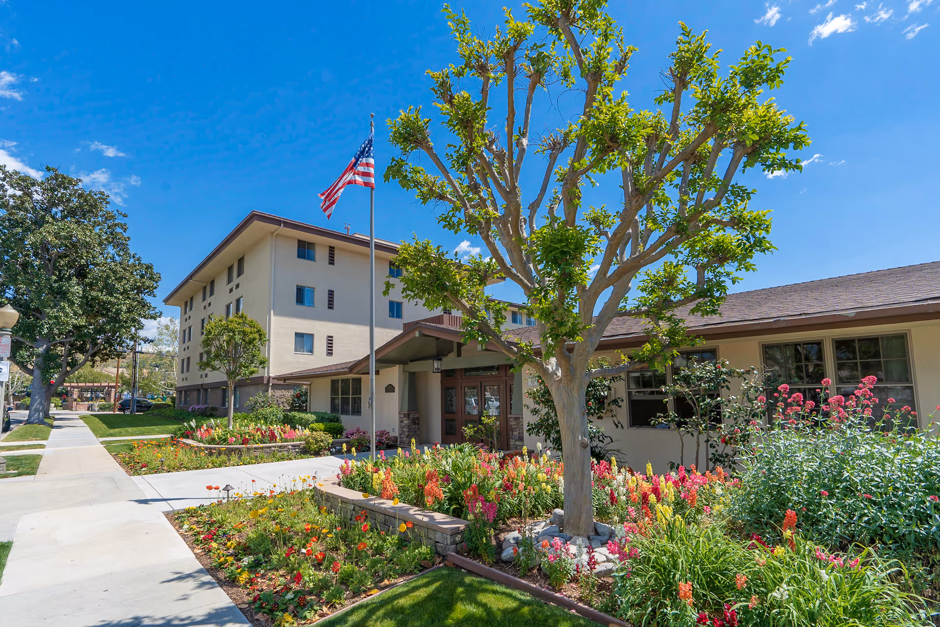 A senior living facility exterior with a colorful flower garden and a tall tree.