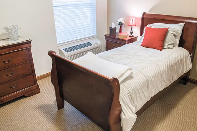 A neatly made single bed with white and beige striped bedding, a red pillow, and a folded white towel at the foot. The bed has a wooden frame. Next to the bed is a wooden nightstand with a red lamp, a tissue box, a small vase with white flowers, and a book. There is a window with closed blinds above a wall-mounted air conditioning unit. On the left side of the image is a wooden dresser with three drawers and a decorative glass item on top. The room has beige carpet and light-colored walls.