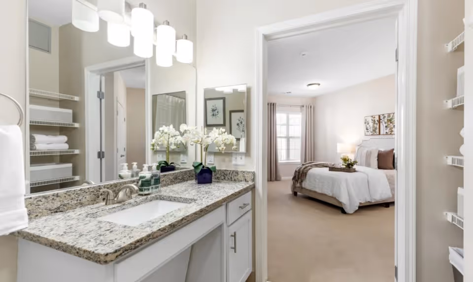 View of a bathroom vanity with a granite countertop, sink, and modern light fixtures above a large mirror. To the right, an open doorway leads to a bedroom with a neatly made bed, beige carpet, and a window with curtains allowing natural light in.