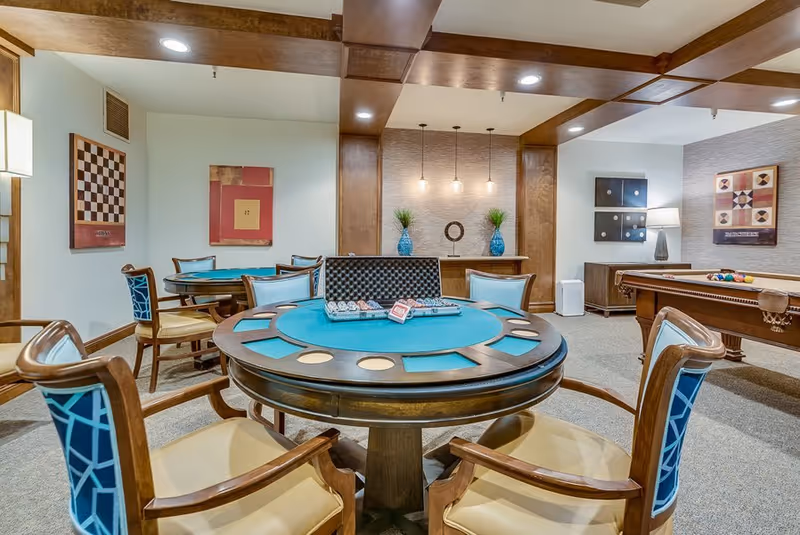 A well-lit game room featuring a round poker table with playing cards and poker chips, surrounded by wooden chairs with blue patterned cushions. In the background, there is a pool table with billiard balls, decorative wall art, and a sideboard with two blue vases and plants under hanging pendant lights. The room has wooden ceiling beams and carpeted flooring.