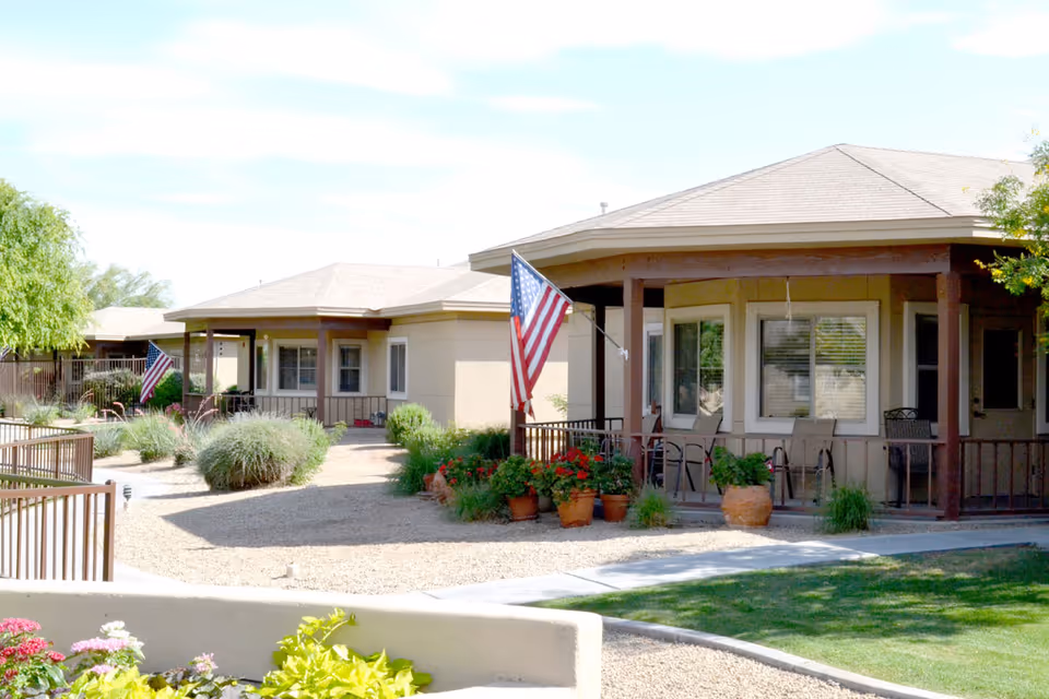 Outdoor view of single-story residential buildings with covered porches, American flags, potted plants, and landscaped gravel and grass areas under a clear sky.
