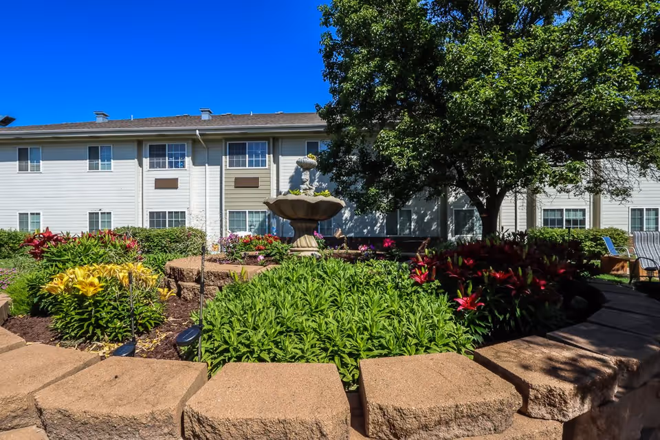 Outdoor garden area at Bailey Pointe at Miracle Hills featuring a stone fountain surrounded by colorful flowers and greenery, with a large tree providing shade and a two-story building in the background under a clear blue sky.