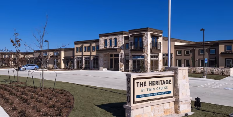 Exterior view of The Heritage at Twin Creeks assisted living and memory care facility with a stone sign in the foreground and a large building in the background under a clear blue sky.