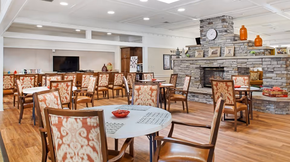 A spacious common area with multiple round tables and patterned chairs arranged around them. The room features a large stone fireplace with a clock and decorative items on the mantel. There are board games on a ledge near the fireplace and a television mounted on the far wall. The flooring is wood, and the ceiling has exposed beams.