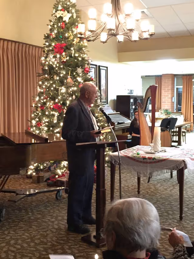 An elderly man stands at a podium speaking into a microphone in a decorated room with a large Christmas tree adorned with lights and ornaments behind him. A woman is seated near a harp in the background, and another elderly person is seated in the foreground. The room has carpeted floors, curtains, and framed pictures on the wall.