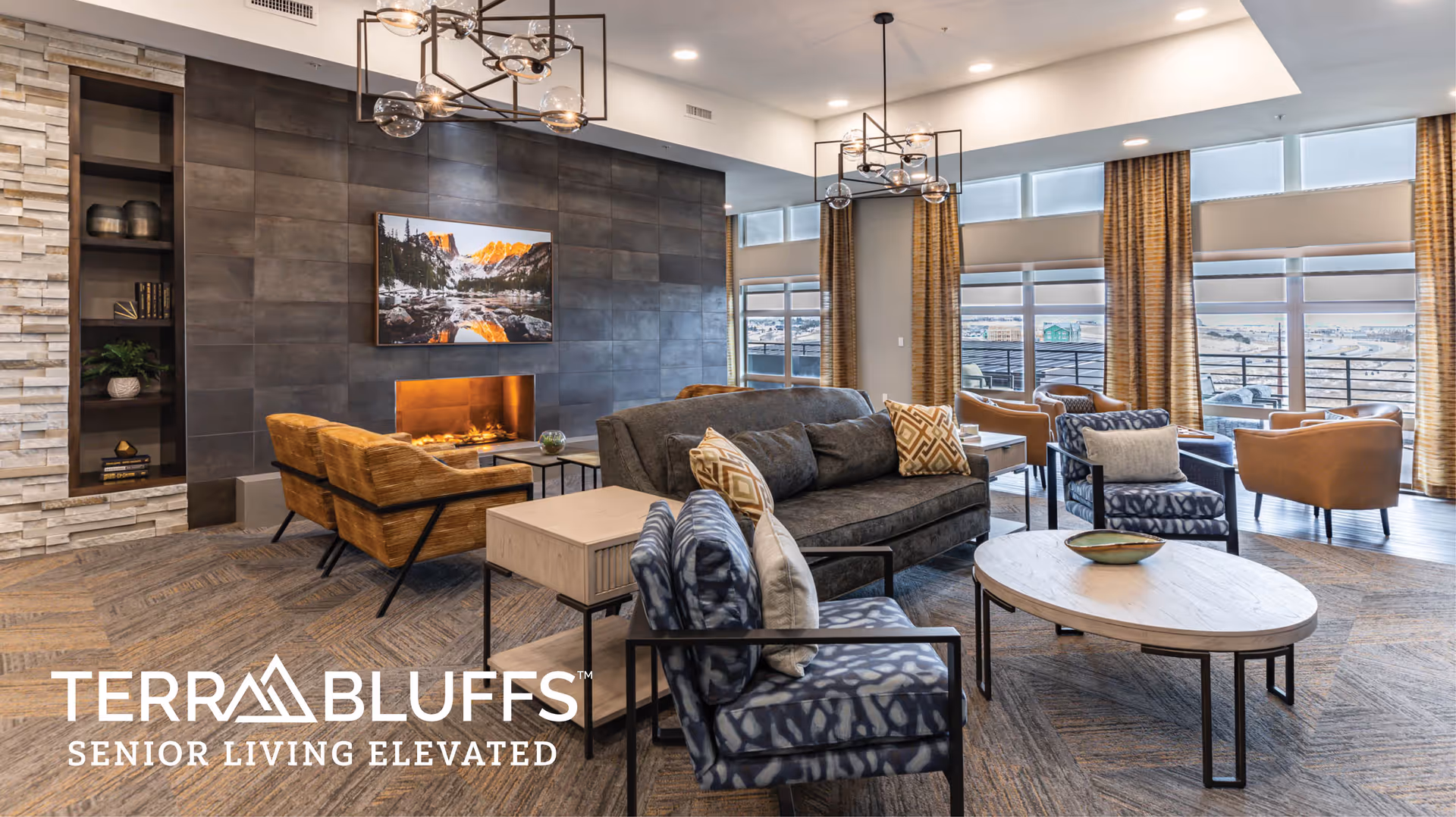 A modern senior living facility lounge area with comfortable seating including sofas and armchairs arranged around a coffee table. The room features a large fireplace with a scenic landscape picture above it, large windows with curtains allowing natural light, and contemporary light fixtures on the ceiling.