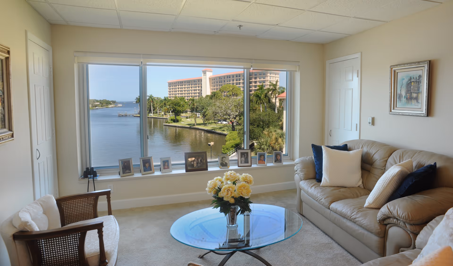 A cozy living room with a large window offering a scenic view of a waterfront and a distant building. The room features a beige leather sofa with white and dark blue pillows, a glass coffee table with a vase of yellow flowers, a wooden chair with a white cushion, and several framed photos on the window sill. The walls are light-colored with a framed painting on one side.
