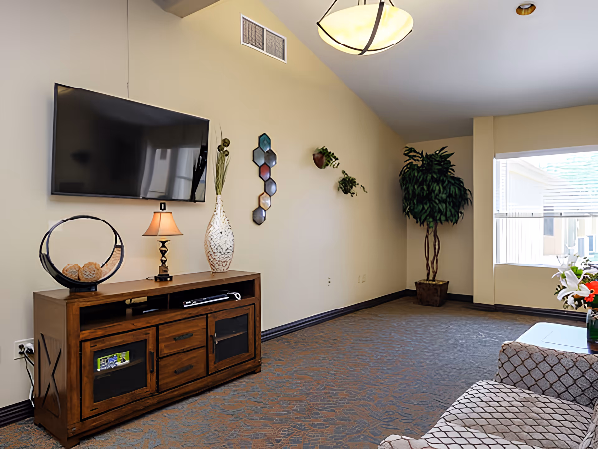 A living room with a mounted flat-screen TV above a wooden media console. The console has decorative items including a lamp, a vase, and a circular sculpture. There are wall-mounted plants and a large potted plant near a window with blinds. A patterned armchair and a small table with flowers are partially visible.