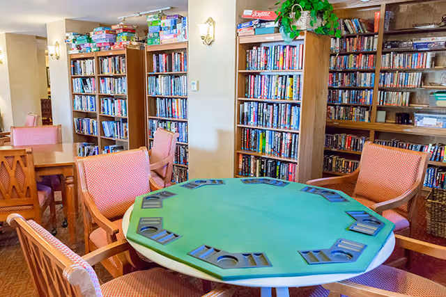 A bright interior room with a green felt octagonal card table surrounded by upholstered chairs and tall bookshelves filled with books and board games.