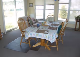 A dining area with a rectangular table covered by a floral tablecloth, surrounded by six wooden chairs. The table is set with placemats, a tissue box, and condiments. The room has large windows letting in natural light and a cushioned armchair near the corner.