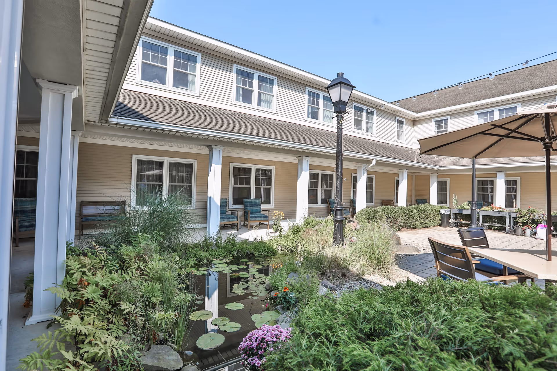 Outdoor courtyard area of a senior living facility featuring a small pond with lily pads, lush greenery, and flowers. Surrounding the courtyard are beige two-story buildings with white columns and multiple windows. There are outdoor seating areas with chairs and tables, including one with a large patio umbrella. A black lamppost stands near the pond under a clear blue sky.