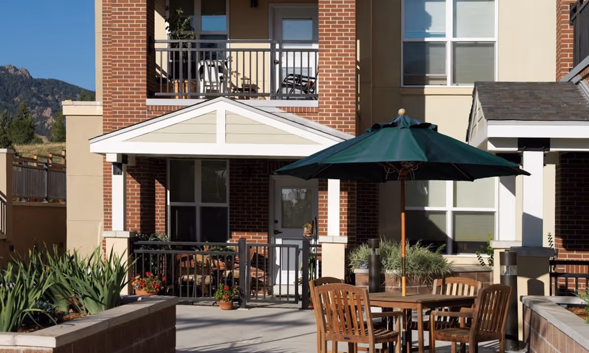 Outdoor patio area at Flatirons Terrace Senior Living featuring a wooden table with four chairs and a large green umbrella. The patio is surrounded by brick and beige building walls with windows and balconies. There are plants and flowers in pots and garden beds around the patio.