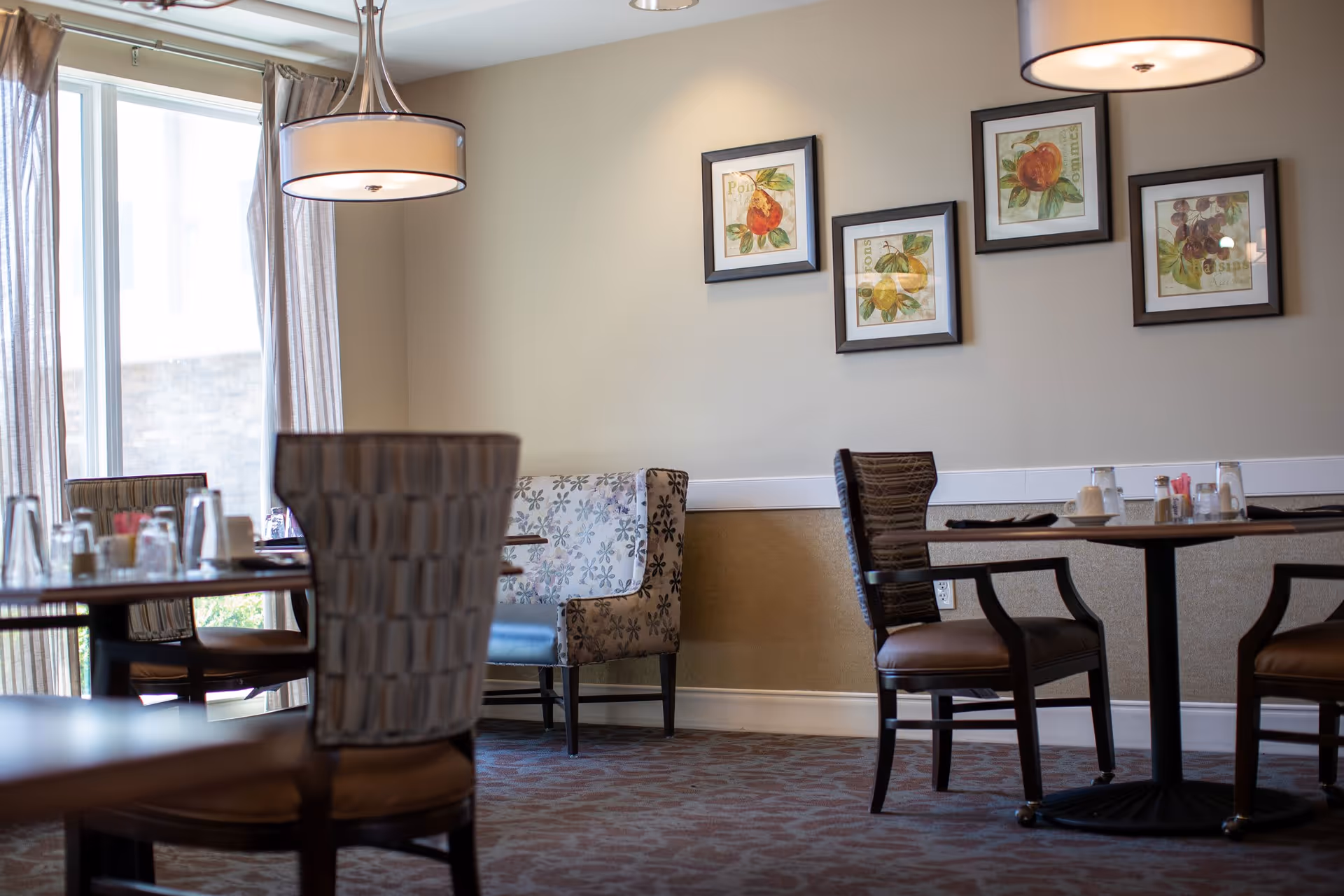 A dining area with several tables and chairs, a large window with curtains allowing natural light, pendant lights hanging from the ceiling, and framed fruit artwork on the beige walls.