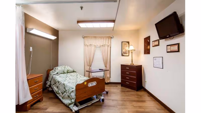 A nursing facility bedroom with a single hospital-style bed covered with a patterned green and white blanket. The room has wooden flooring, a wooden nightstand with drawers on the left, a small overbed table, a chest of drawers with a lamp on top, and a wall-mounted TV. The window is covered with beige curtains, and several framed pictures hang on the walls.
