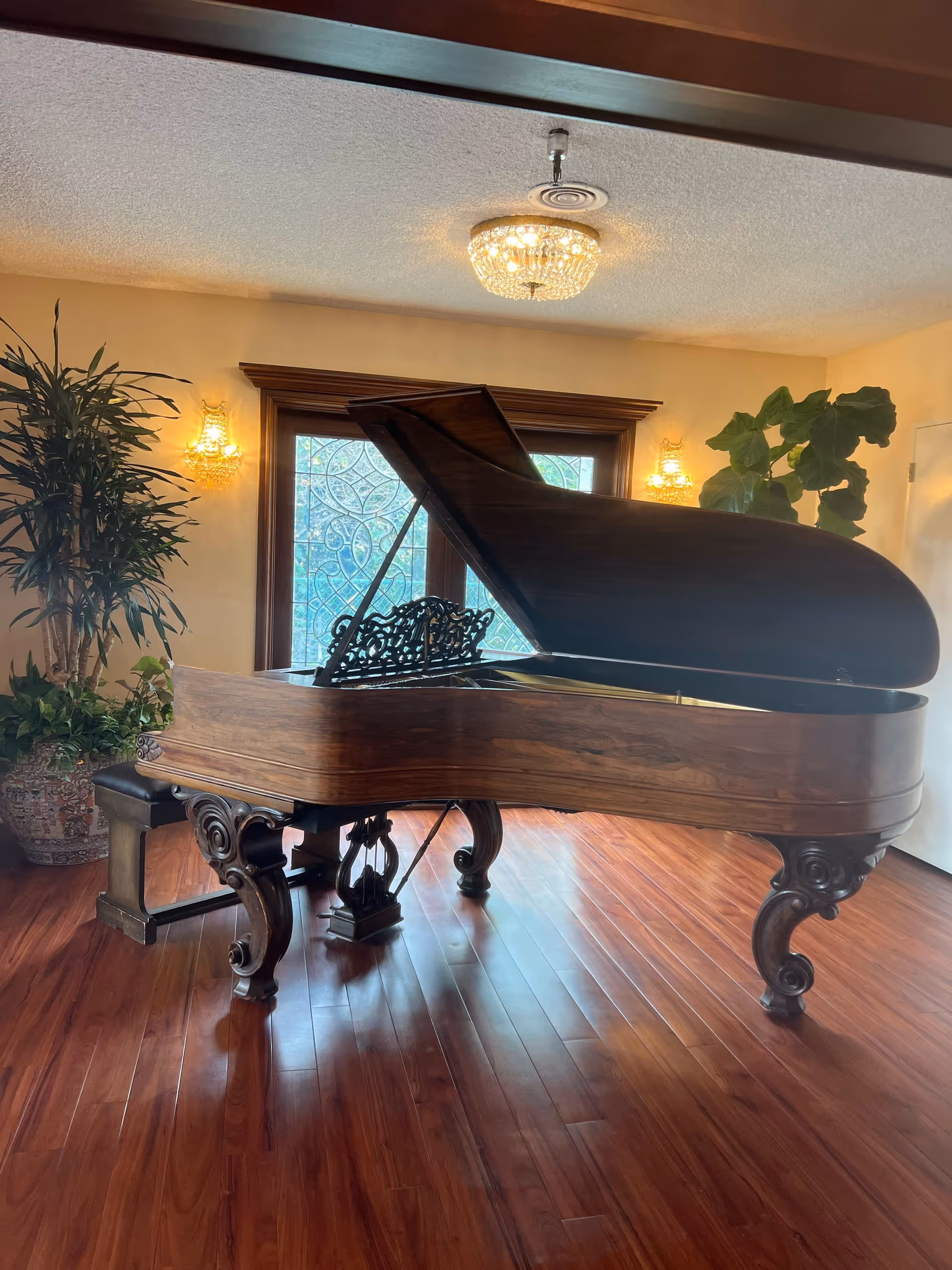 A grand piano with ornate carved legs sits on polished wooden flooring in a room with beige walls. Behind the piano are two large windows with decorative glass panels, flanked by two wall sconces with crystal-like designs. There are large potted plants on either side of the piano, and a crystal chandelier hangs from the ceiling.