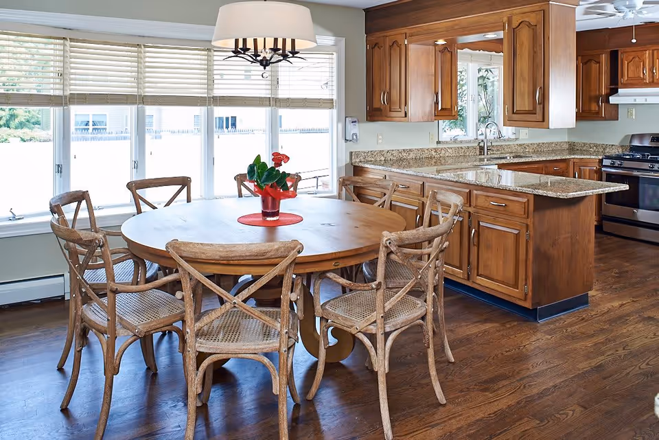 A bright kitchen and dining area with a round wooden table surrounded by six rustic wooden chairs. The kitchen features wooden cabinets, granite countertops, a stainless steel stove, and a large window with blinds letting in natural light. A small red flower arrangement is centered on the dining table.
