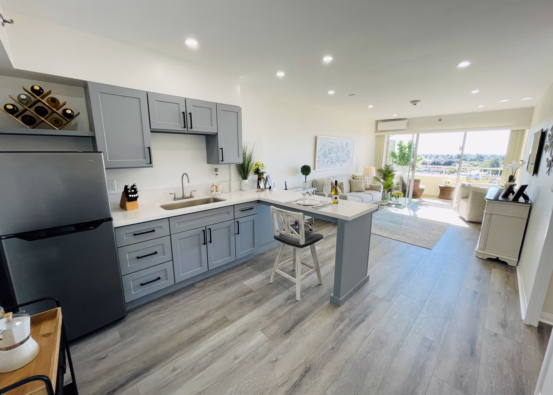 Bright and modern open-concept living space featuring a kitchen with gray cabinets, a refrigerator, a sink, and a small island with a chair. The living room area has a sofa, coffee table, plants, and large sliding glass doors leading to a balcony with outdoor seating and a view of greenery and buildings in the distance.