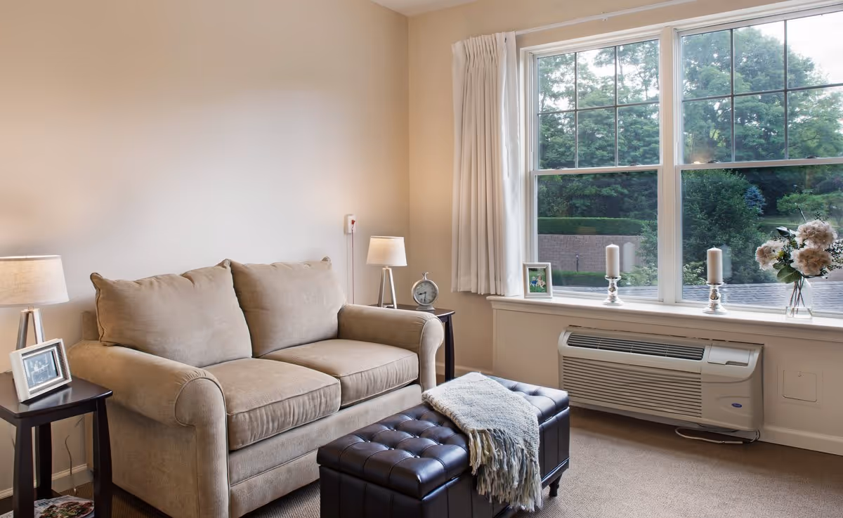 Bright living room with a beige loveseat, dark ottoman, side tables and lamps, and a large window overlooking greenery.