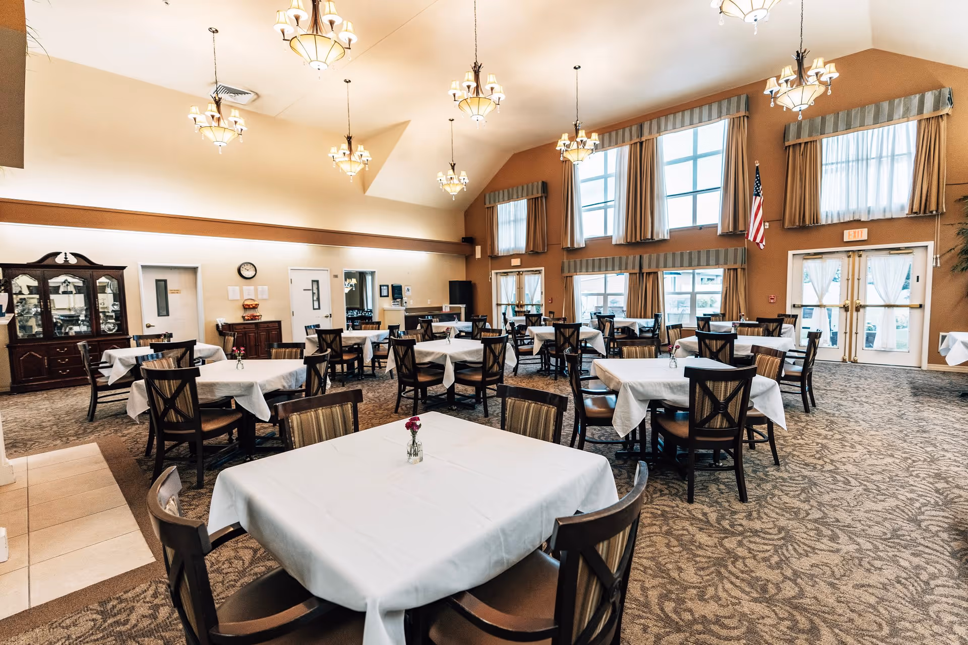 A spacious dining room with multiple tables covered in white tablecloths, each surrounded by wooden chairs. The room has high ceilings with several hanging light fixtures, large windows with curtains, and an American flag near the exit doors. There is a carpeted floor with a patterned design and a wooden cabinet against one wall.