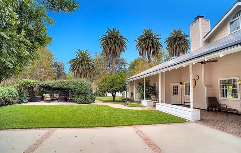 Outdoor view of a senior living facility showing a well-maintained lawn, palm trees, and a covered patio area with seating. The building has a light-colored exterior with a chimney and ceiling fans under the porch roof.