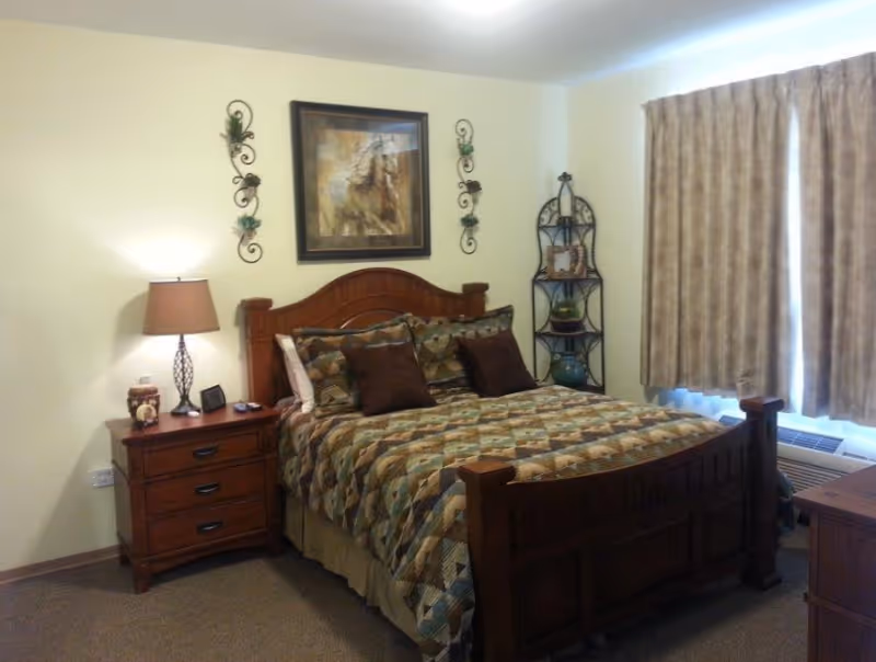 A cozy bedroom with a wooden bed frame and a patterned quilt. There are two dark brown pillows and several patterned pillows on the bed. A wooden nightstand with a lamp, clock, and small decorative items is beside the bed. Above the bed hangs a framed picture of a horse, flanked by two decorative wall sconces. A window with beige curtains and an air conditioning unit below it is on the right side of the room. A metal corner shelf with decorative items is also visible.