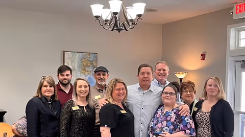 A group of nine adults standing closely together indoors, smiling at the camera. The room has neutral-colored walls, a chandelier overhead, a piece of wall art, and a lamp in the corner. There is an exit sign above a door in the background.