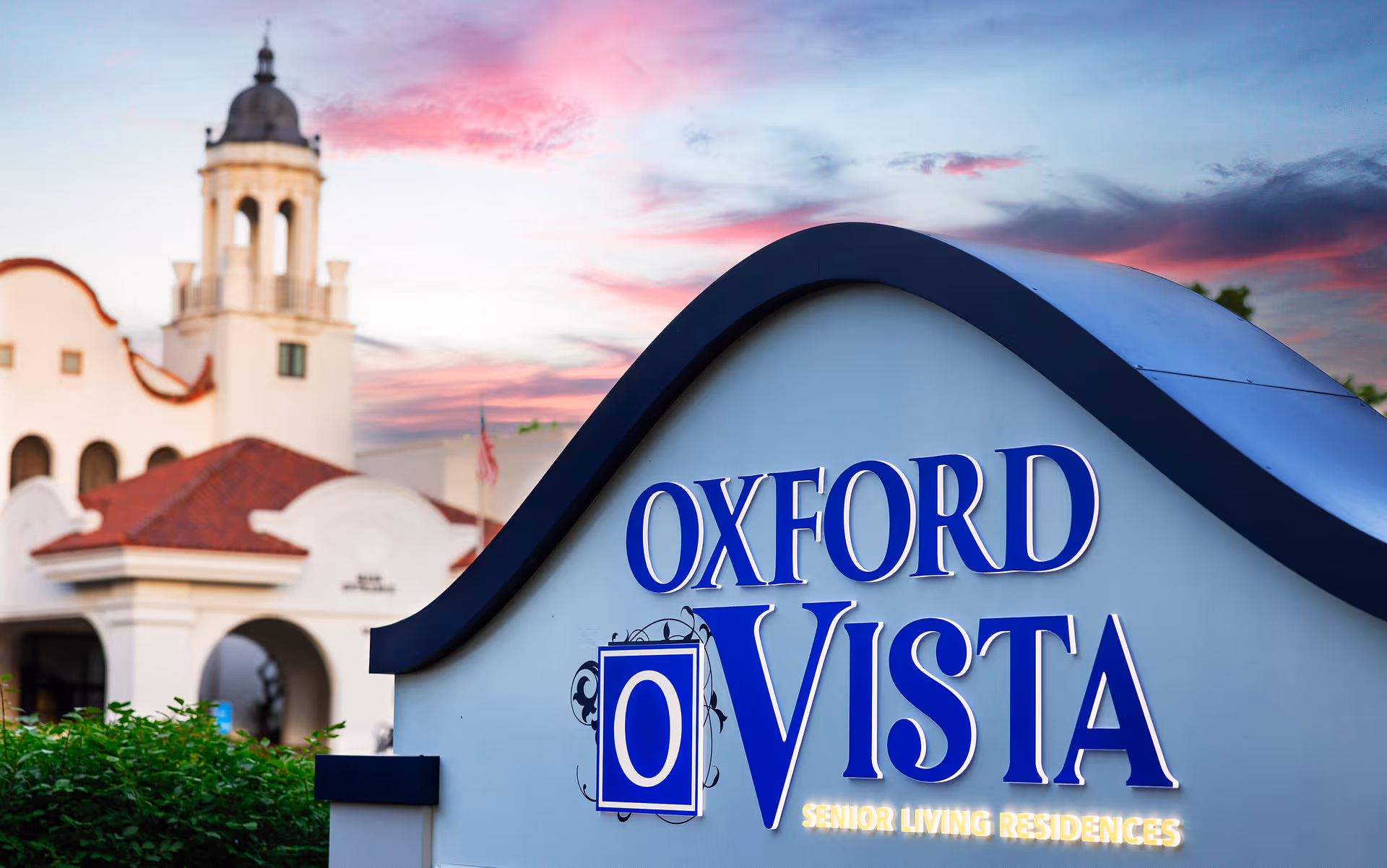 Sign for Oxford Vista Senior Living Residences with a white building featuring a tower and red-tiled roof in the background under a colorful sunset sky.