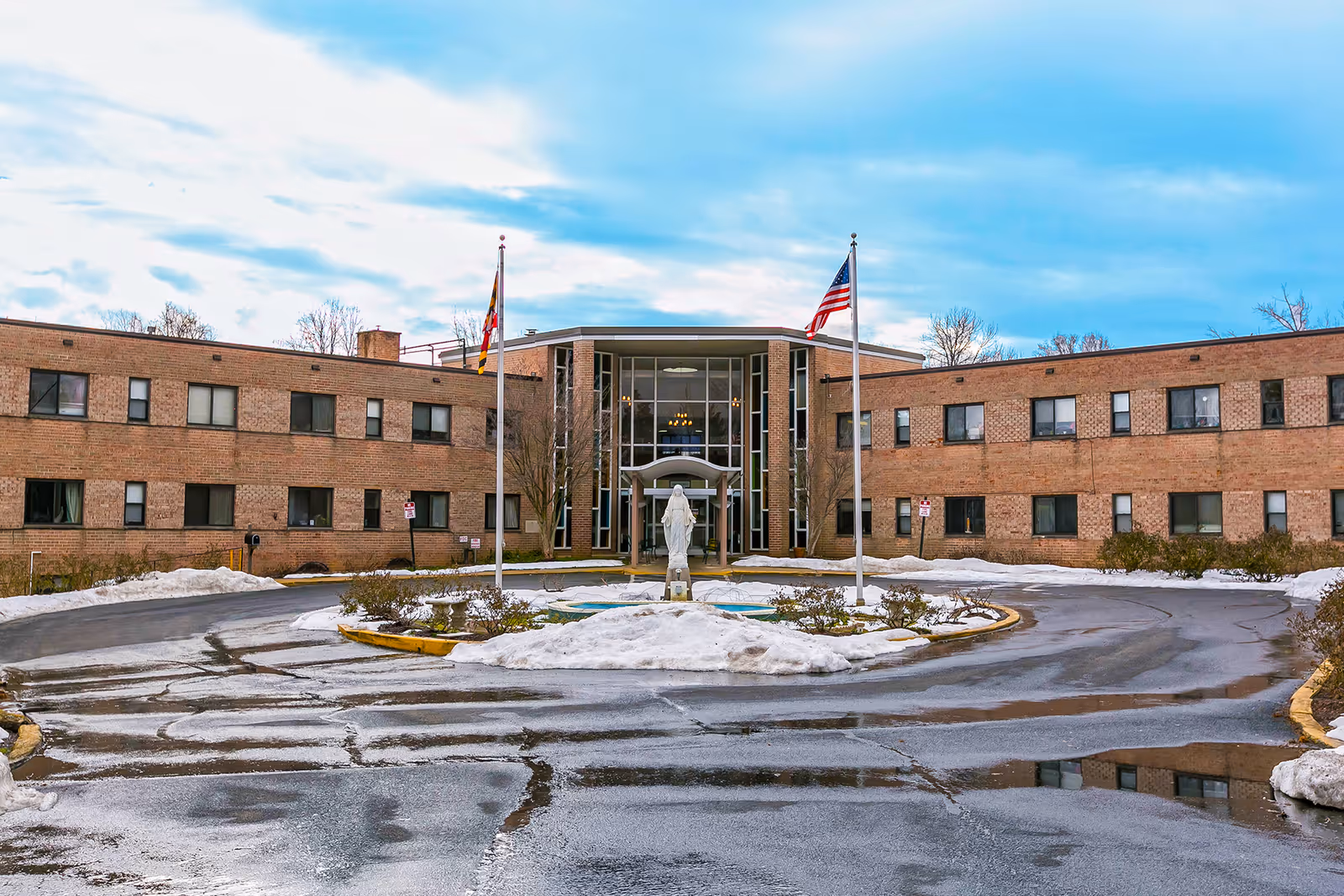Front exterior view of Villa Rosa Nursing and Rehabilitation building with a circular driveway, two flagpoles with flags, a statue in the center, and patches of snow on the ground.