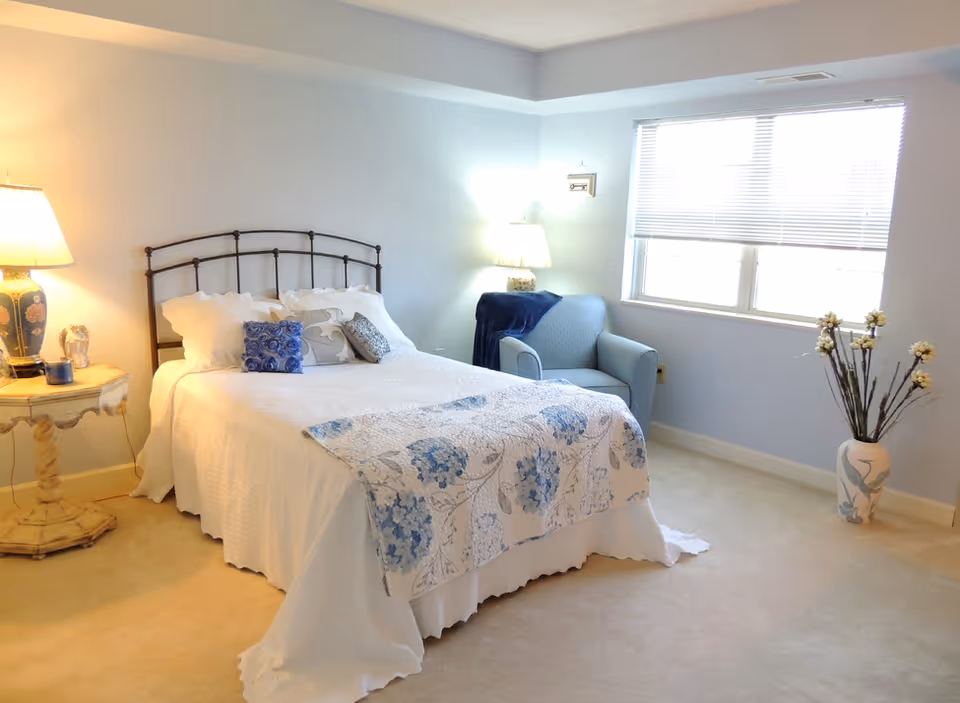 A bright bedroom with a neatly made bed featuring white and blue floral bedding, a blue armchair with a dark blue throw blanket, a wooden side table with a lamp and decorative items, a large window with blinds, and a vase with tall flowers on the floor.