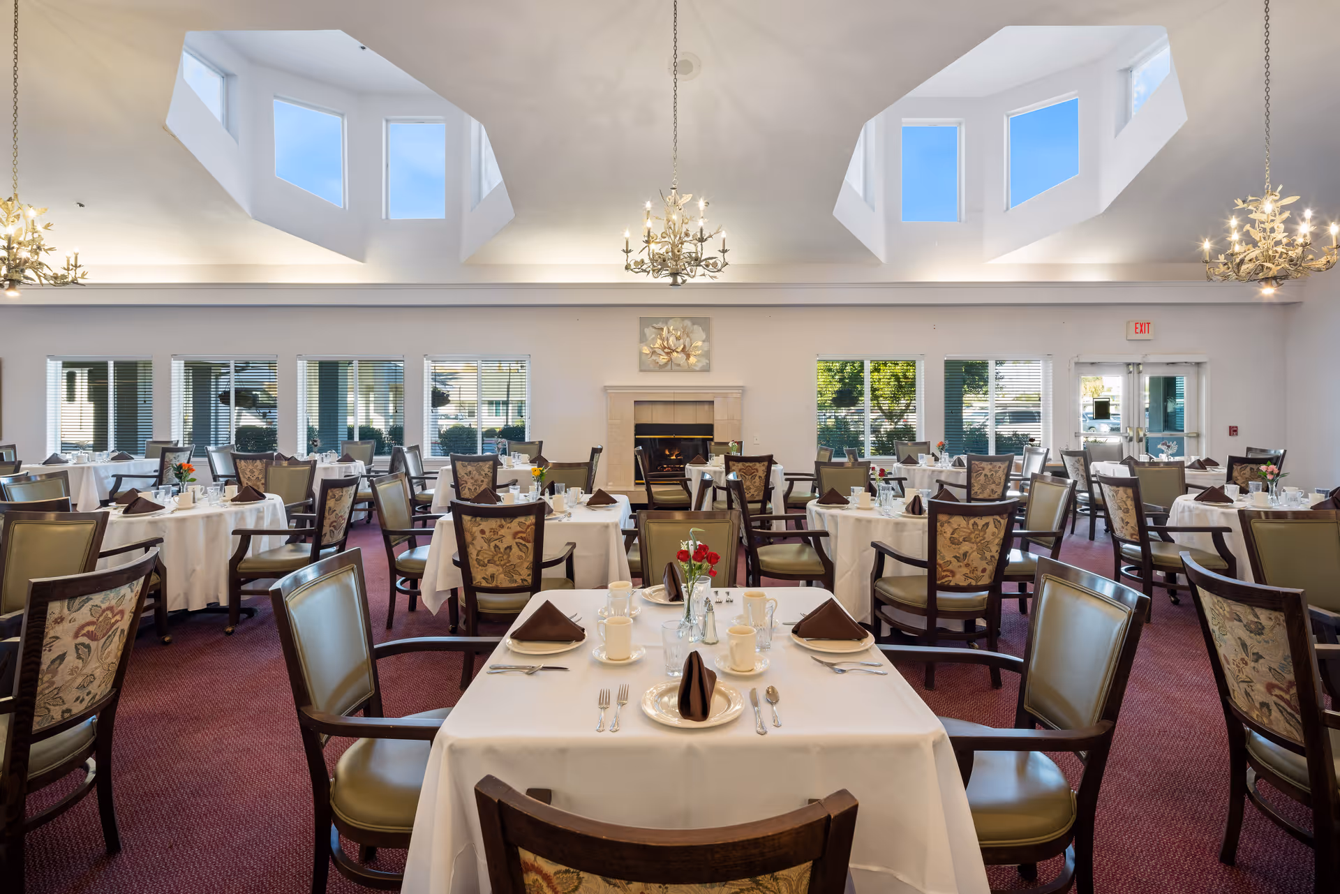 A spacious dining room with multiple round tables covered in white tablecloths, each set with plates, cups, silverware, and brown folded napkins. The chairs have patterned upholstery and green leather seats. The room features large windows, a fireplace in the center, and three ornate chandeliers hanging from a high ceiling with skylights.