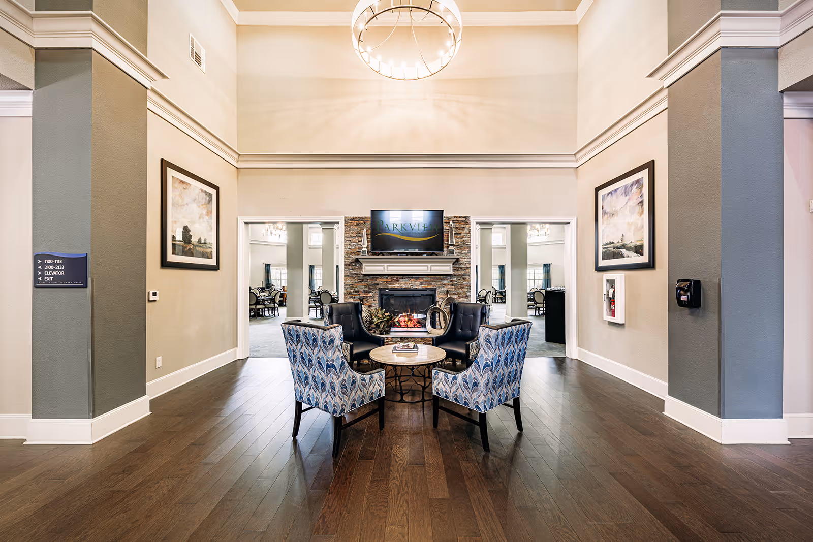 A spacious and well-lit common area in a senior living facility featuring a seating arrangement with four chairs around a small round table in front of a stone fireplace. Above the fireplace is a TV screen displaying the text 'Parkview on Hollybrook'. The room has high ceilings with crown molding, two framed landscape paintings on the walls, and hardwood flooring. Two open doorways on either side of the fireplace lead to a dining area with tables and chairs.